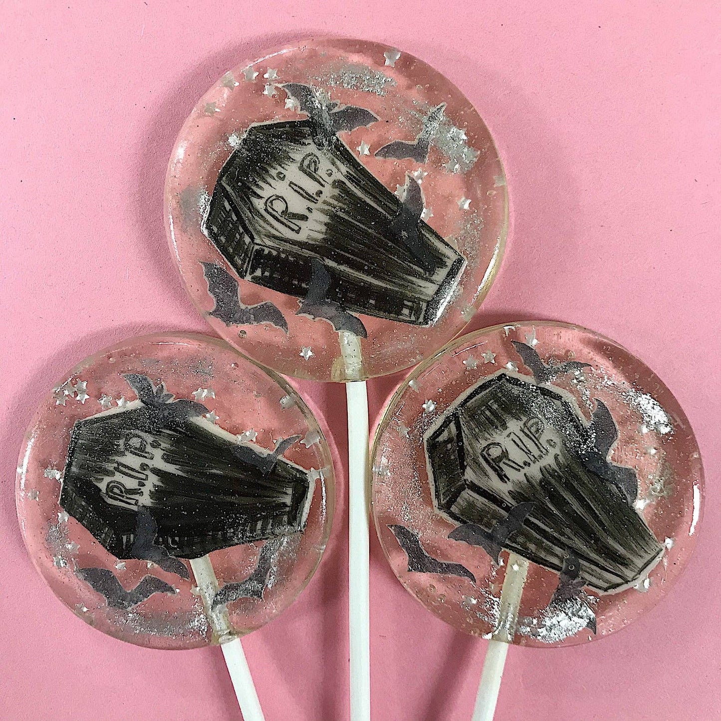 Round lollipops with black fondant coffin designs, silver glitter stars, and wafer paper bats on a pink background Peace Coffin Lollipops