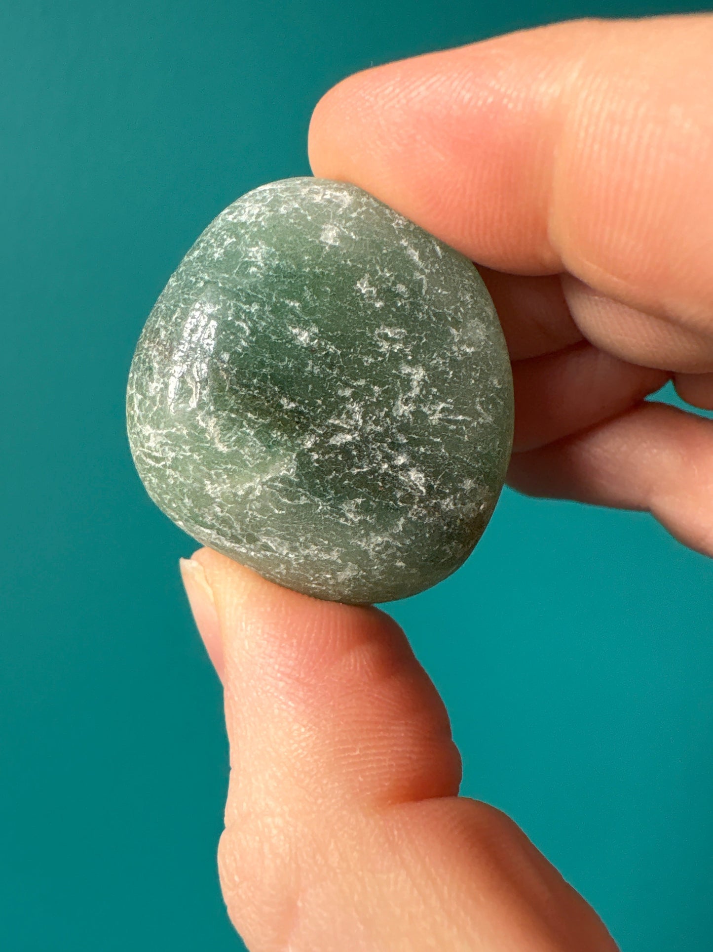 Green aventurine tumble stone held between fingers on teal background