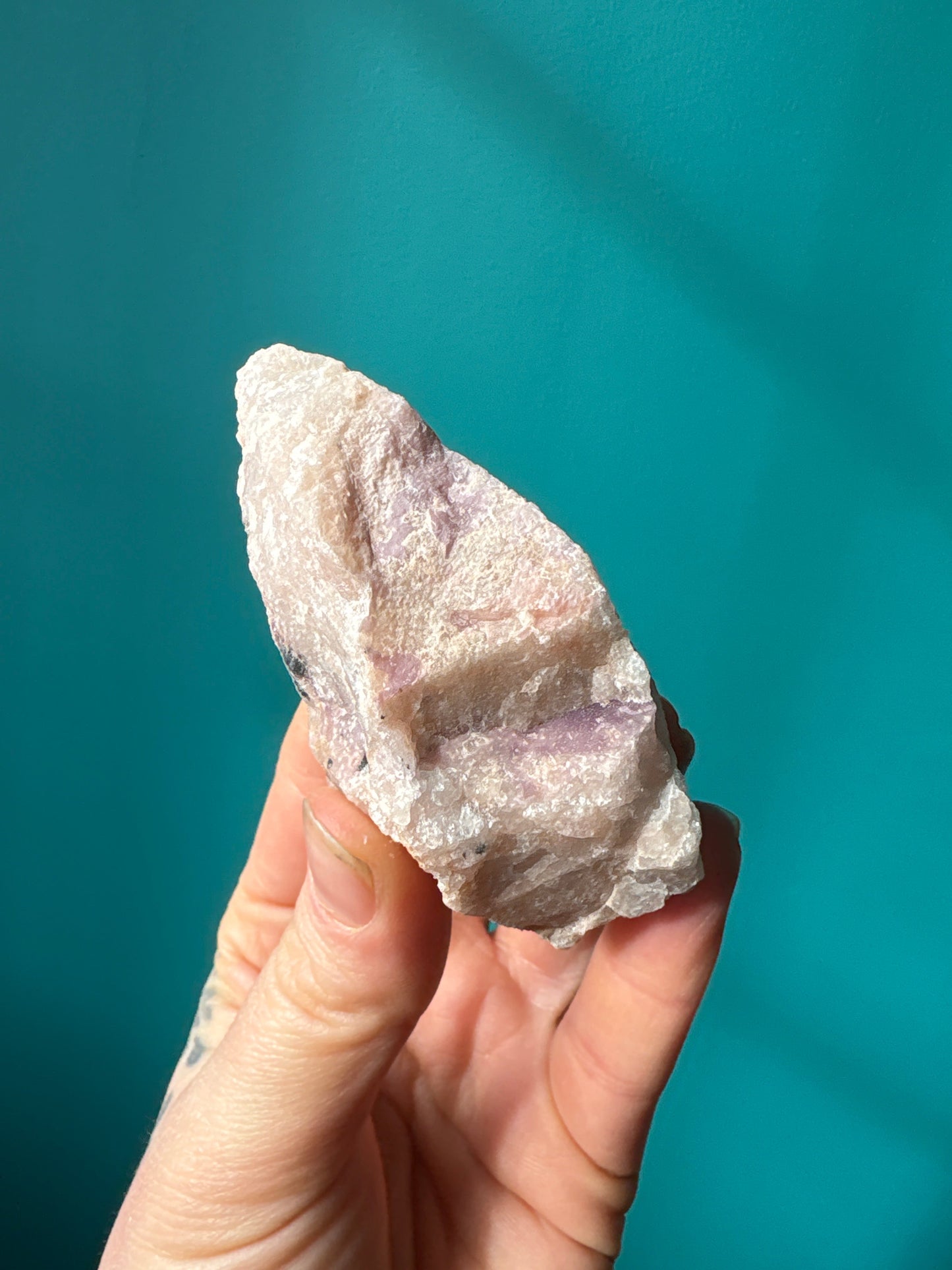 hand holding a raw Pink Amblygonite gemstone with pale pink and white hues against a blue background