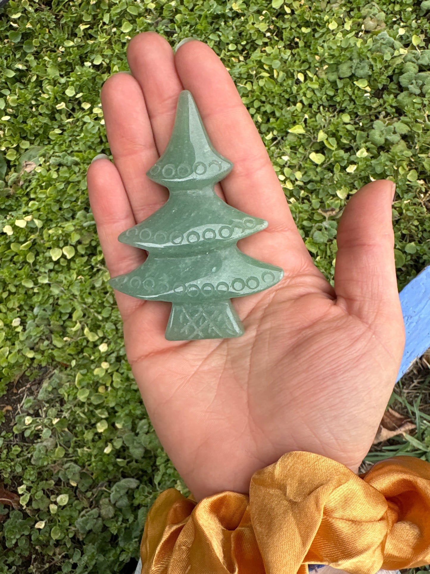 Hand holding a hand carved green aventurine christmas tree crystal with engraved patterns against a green leafy background