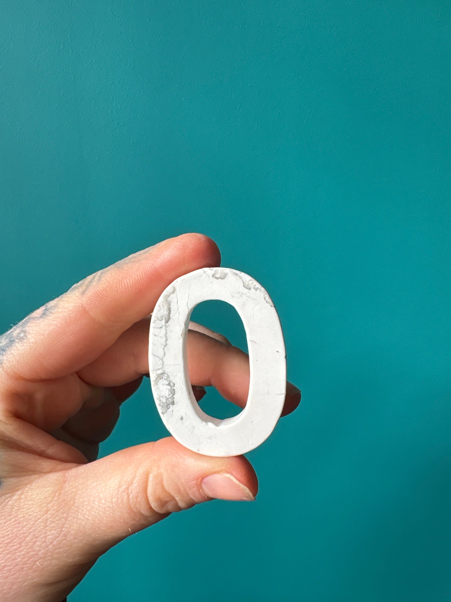 Hand holding a howlite stone carved into the number zero against a teal background, symbolizing positivity and new beginnings.
