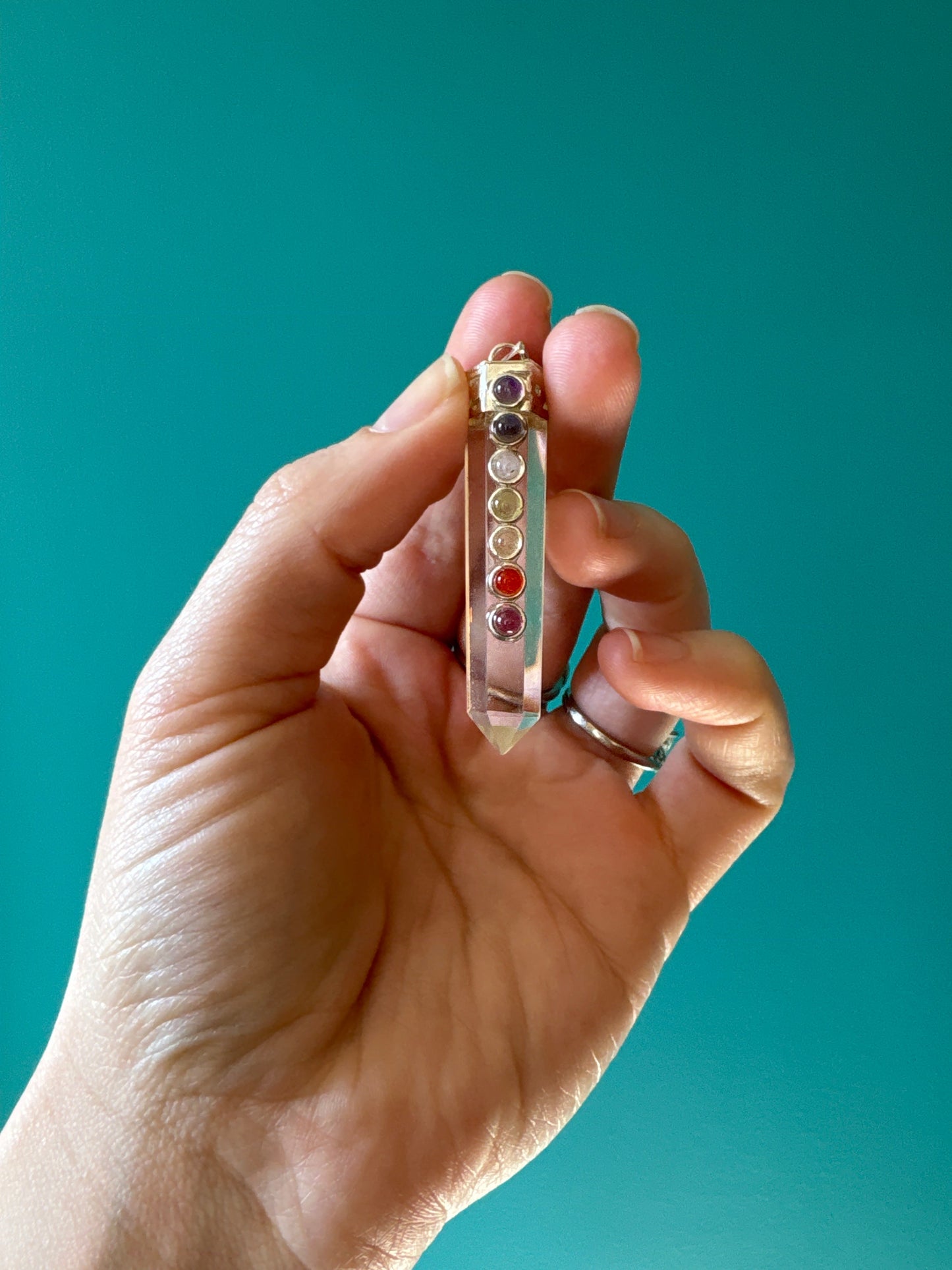 Hand holding a clear quartz polished chakra pendant with seven chakra stones against a teal background