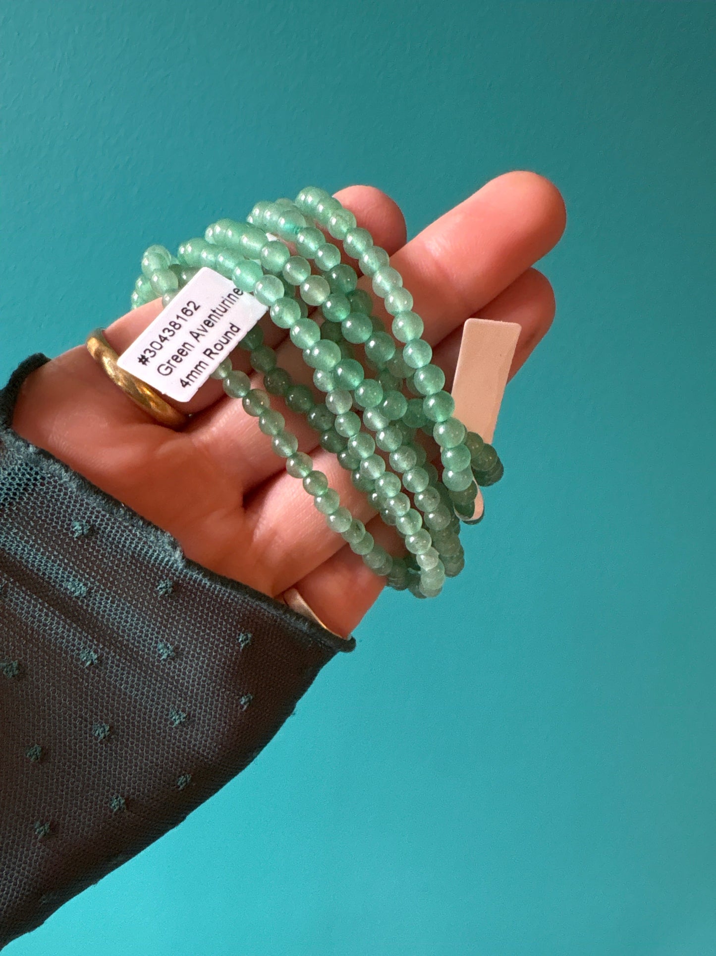 Hand holding multiple strands of 4mm green aventurine bead bracelets against a teal background