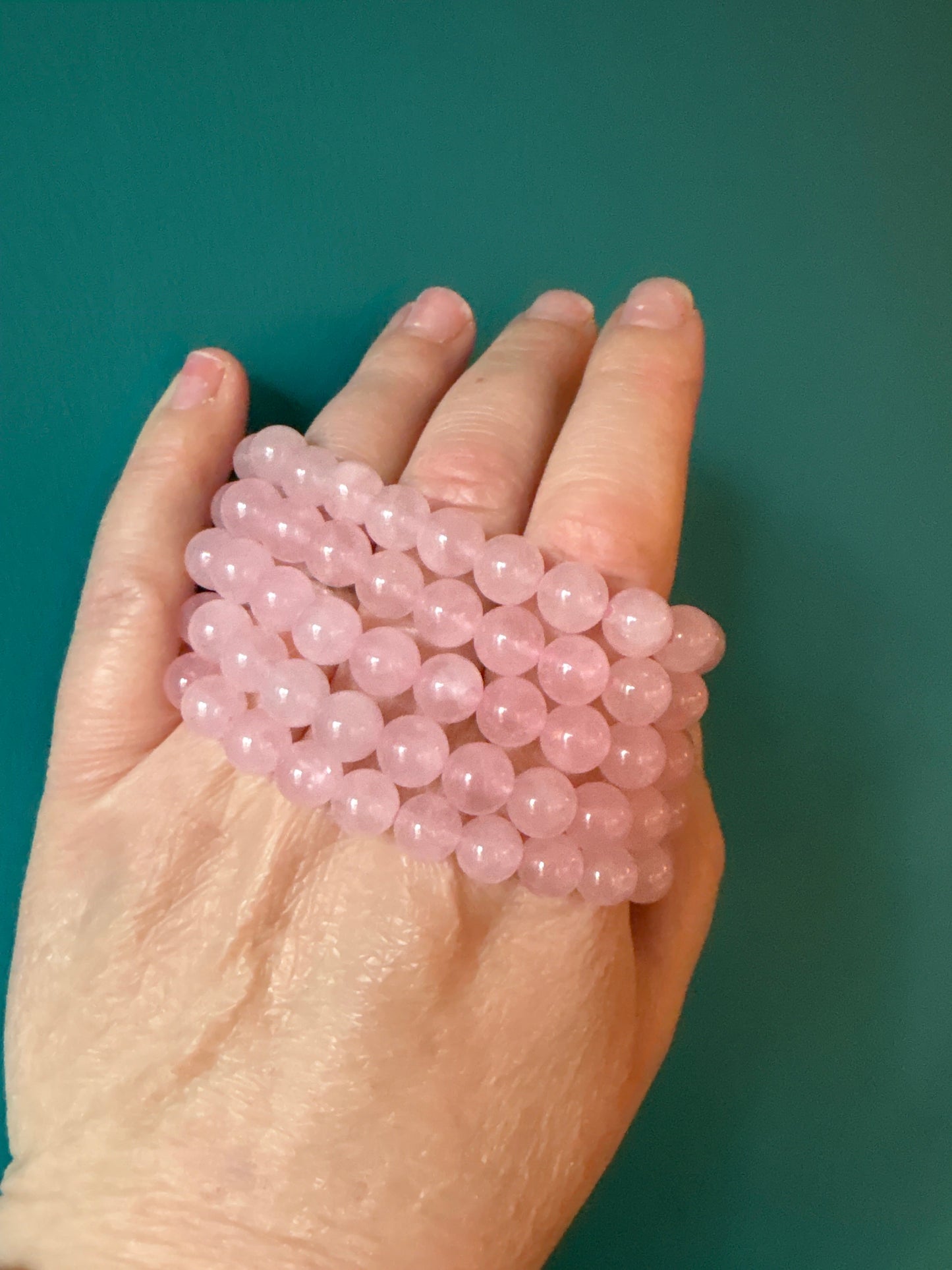 Hand holding multiple strands of polished 8mm rose quartz bead bracelets on a teal background