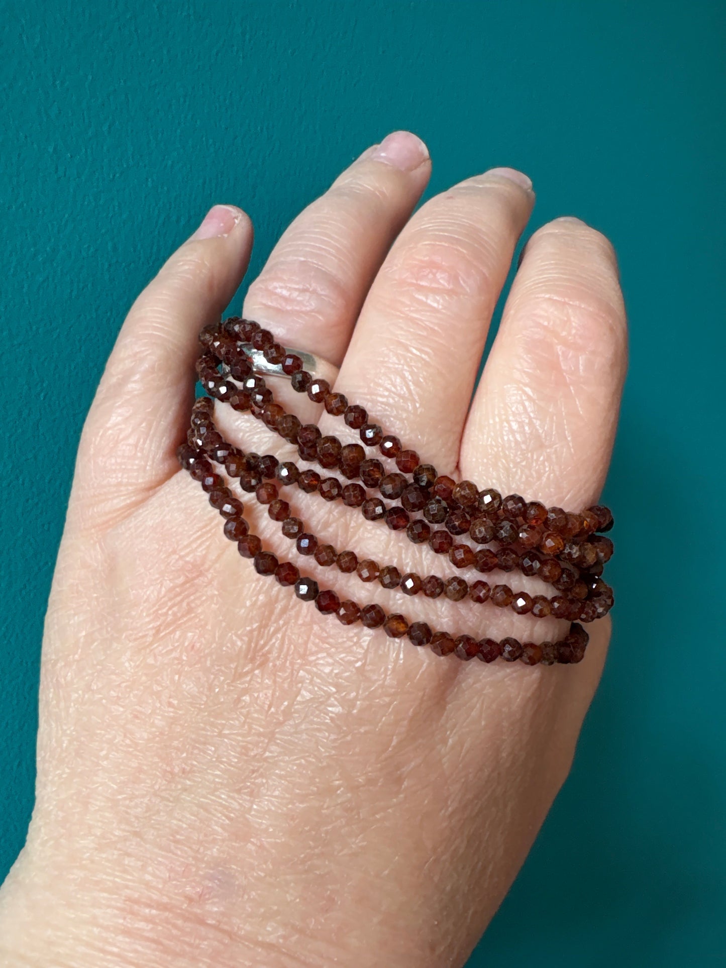 Close-up of hand wearing multiple strands of faceted 4mm orange garnet bead bracelet against teal background