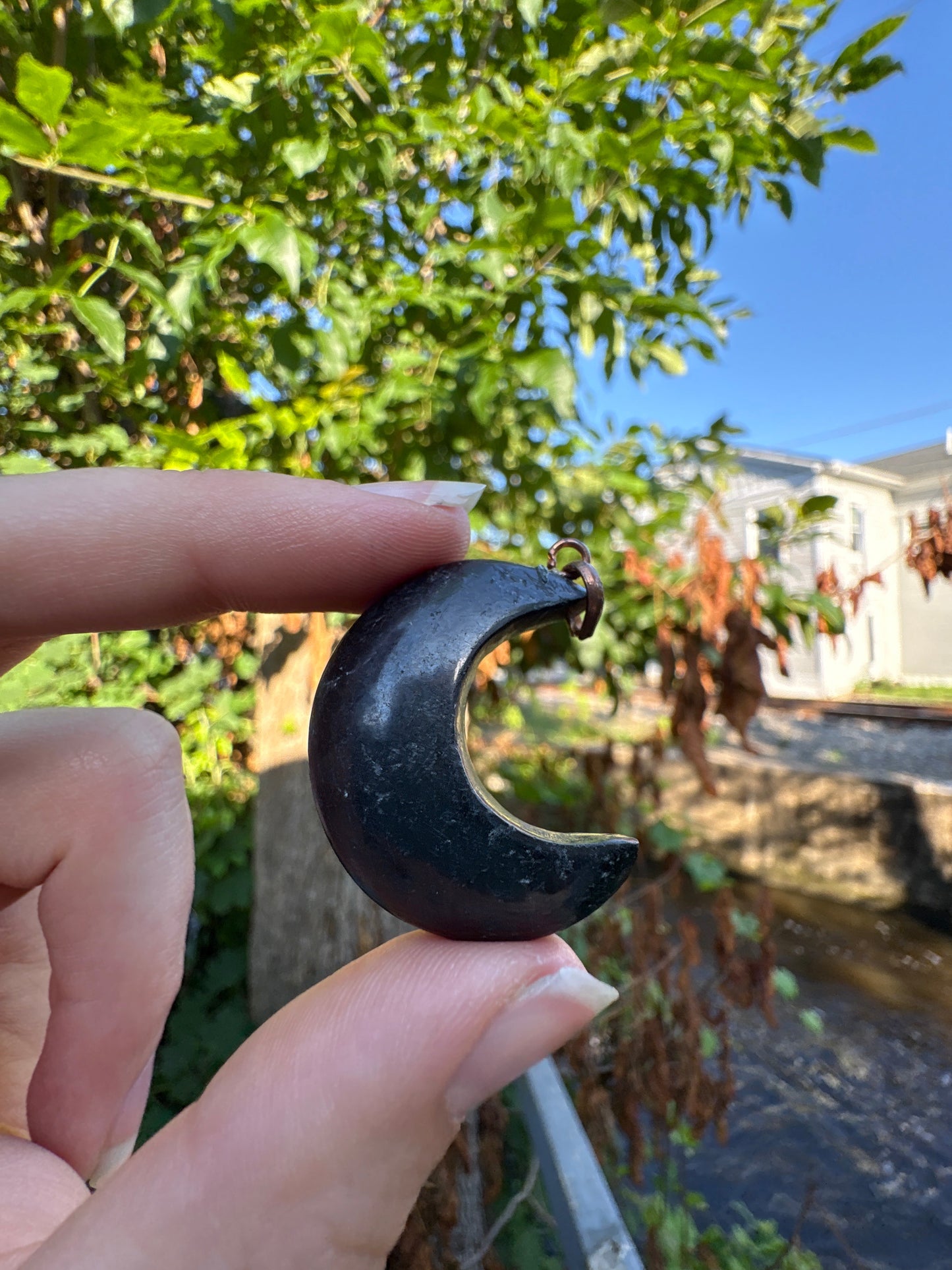 Hand holding a crescent-shaped Shungite Moon Pendant outdoors with trees and buildings in the background
