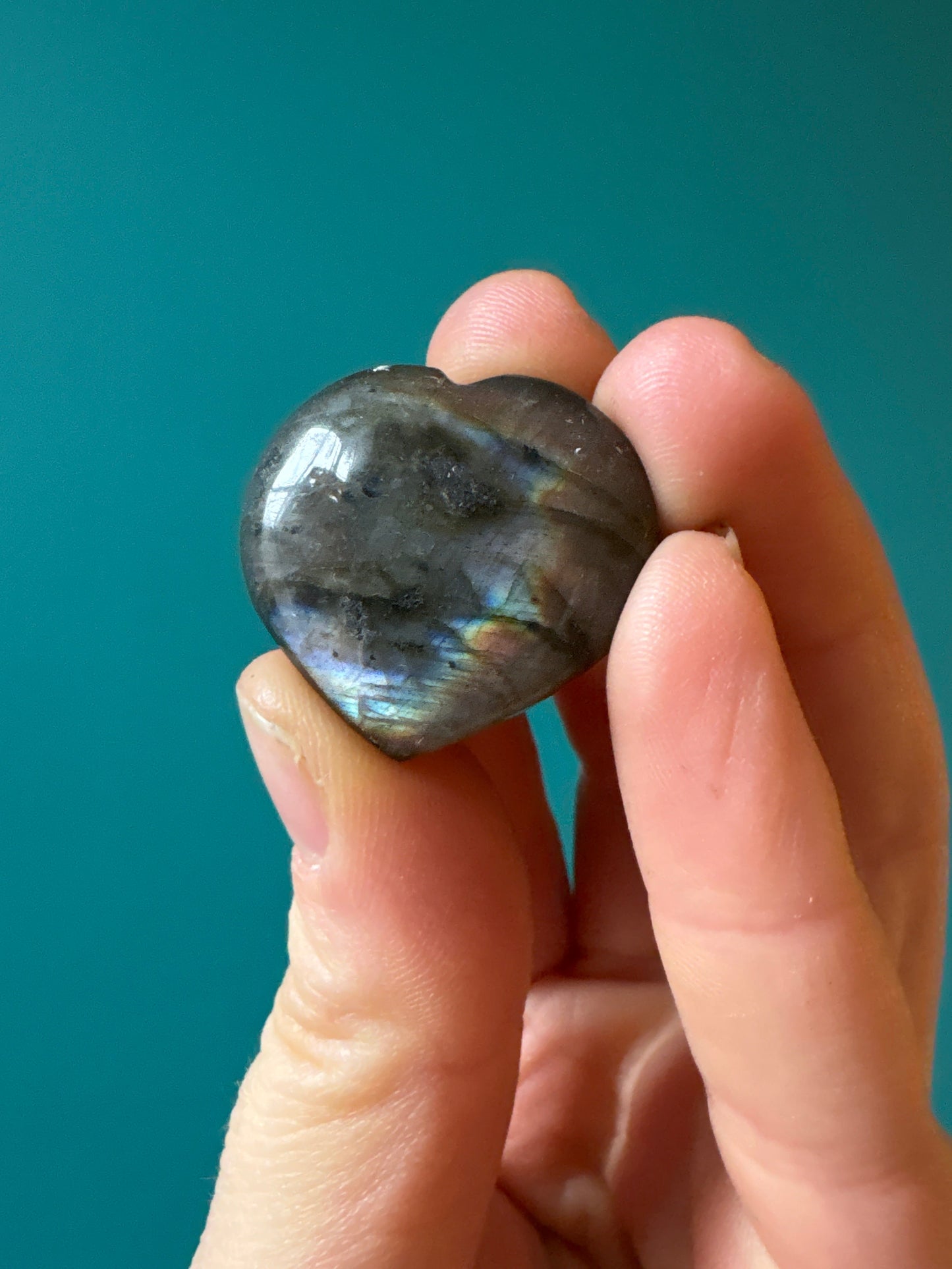 Hand holding a polished heart-shaped labradorite gemstone with iridescent flashes against a teal background