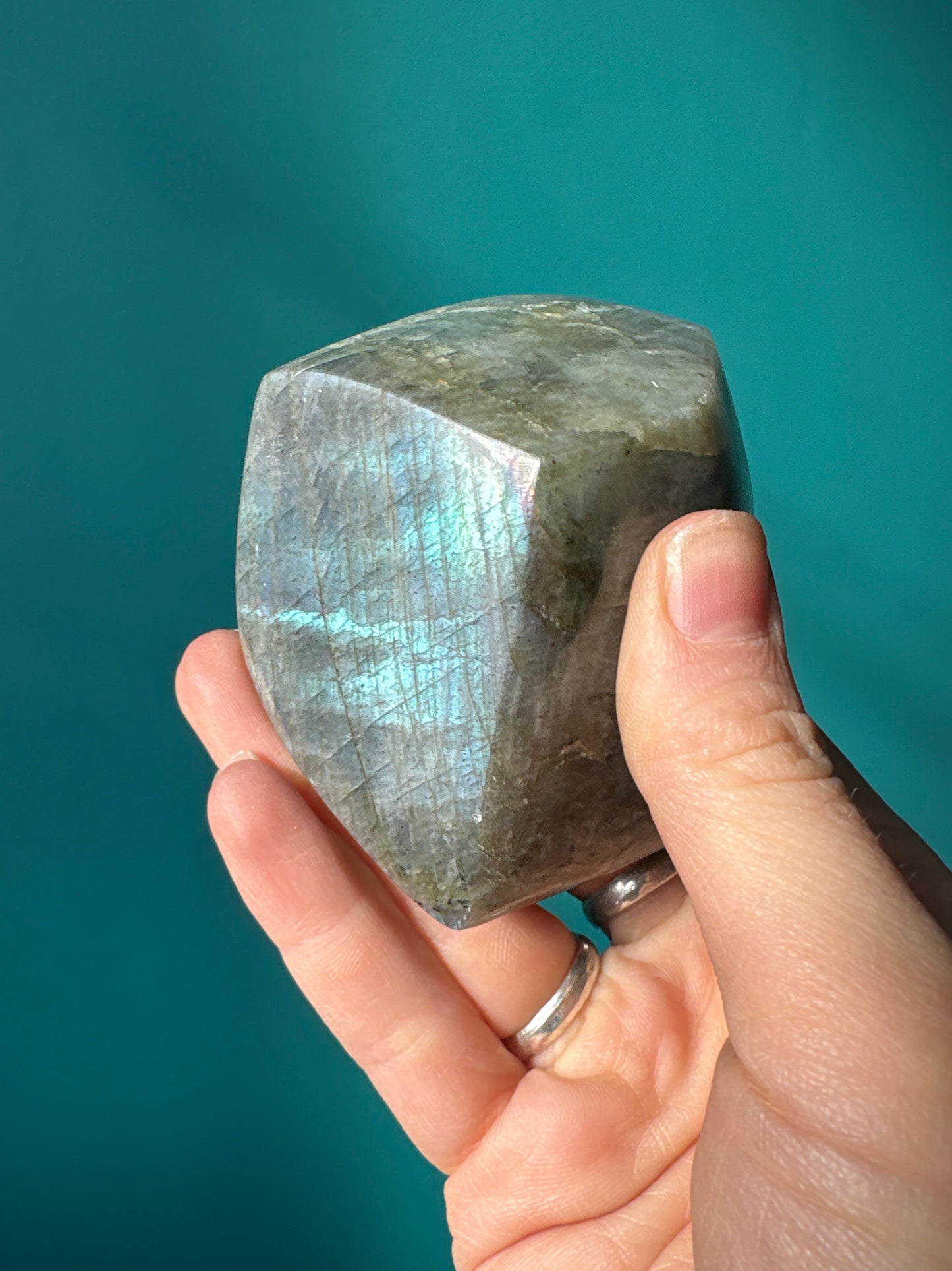 Hand holding a polished Labradorite Freeform stone showing its iridescent blue and green flashes against a teal background