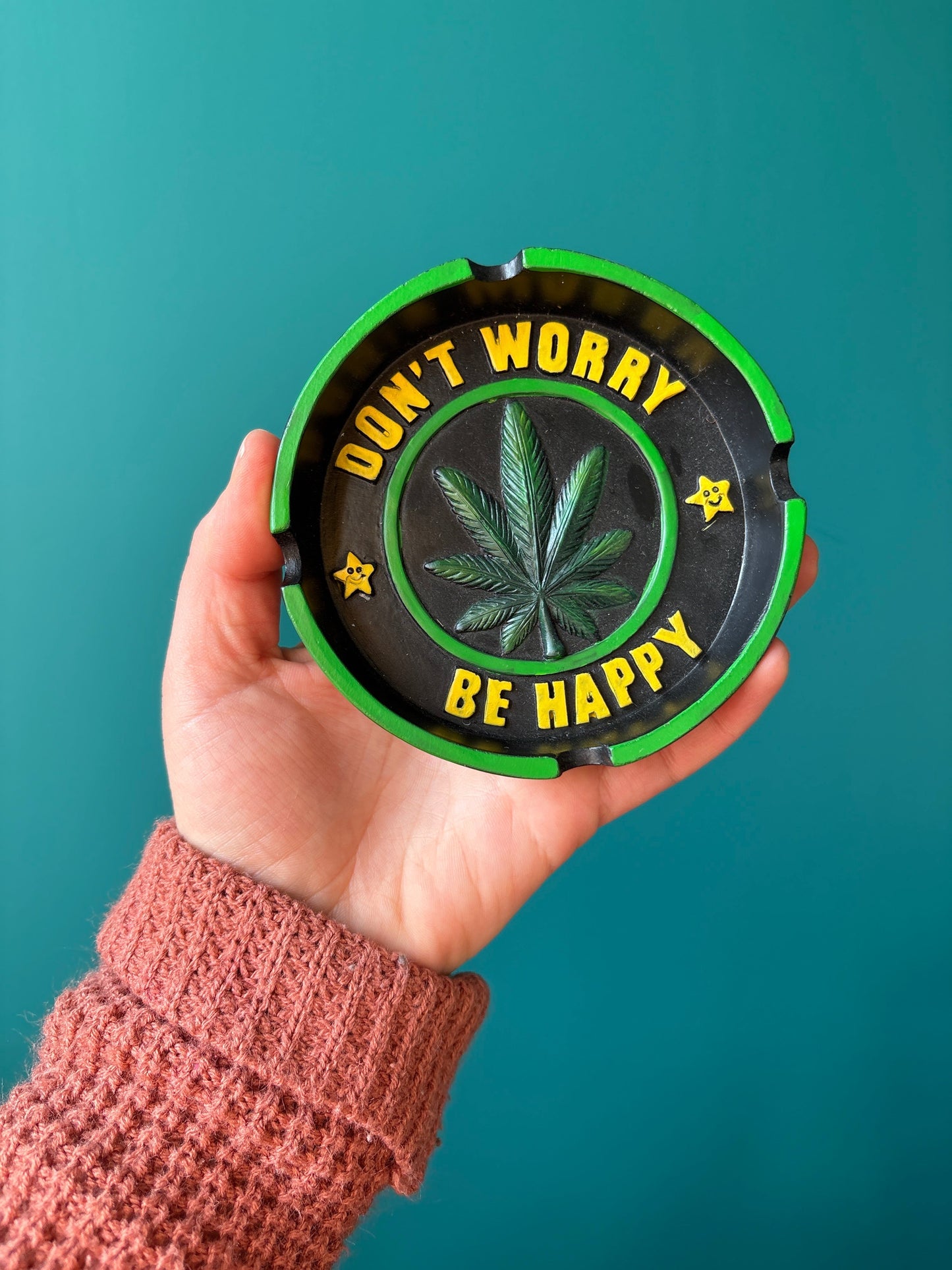 hand holding a green and black worry be happy ashtray with a cannabis leaf and the text "Don't worry be happy"