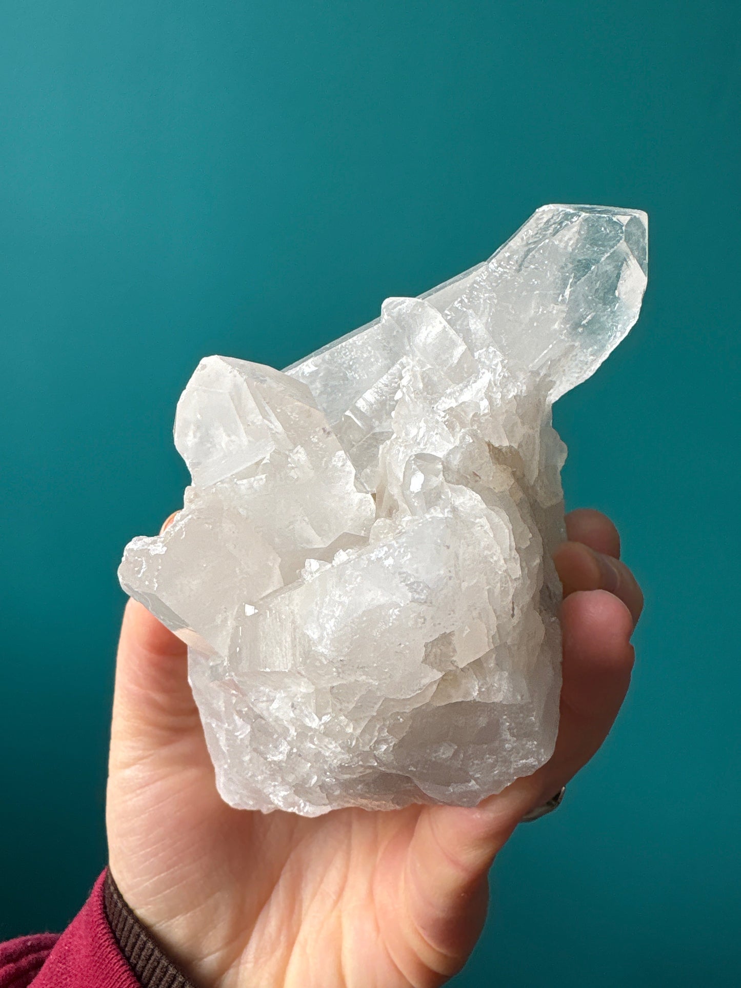 Clear quartz cluster crystal from Brazil held in hand against teal background