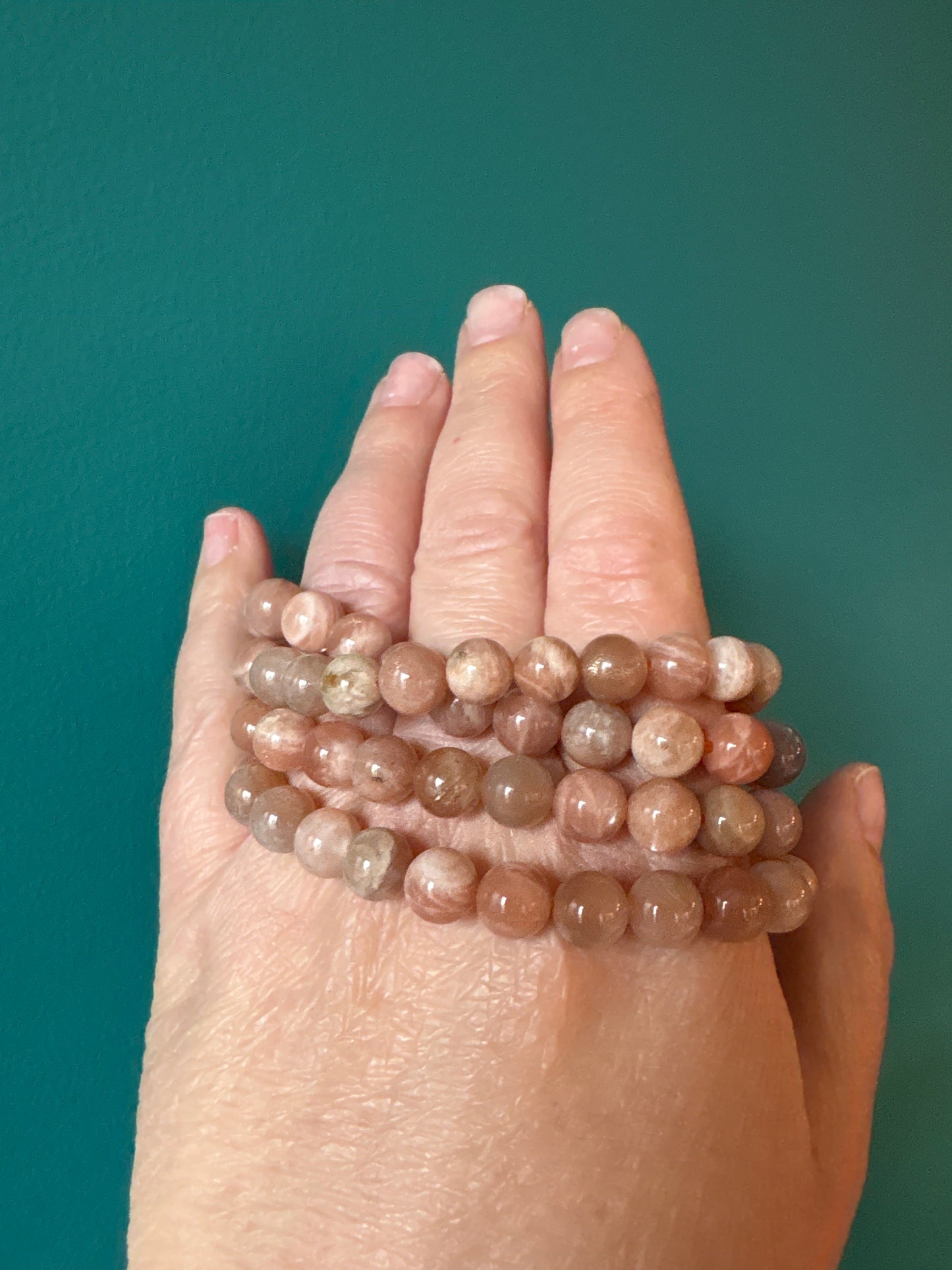 Hand wearing multiple strands of 8mm Sunstone Bracelet beads against a teal background