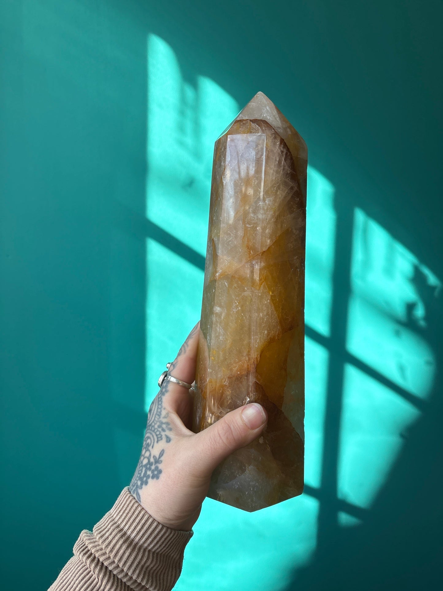 Hand holding a large Tower Golden Healer crystal with teal background and sunlight shadows