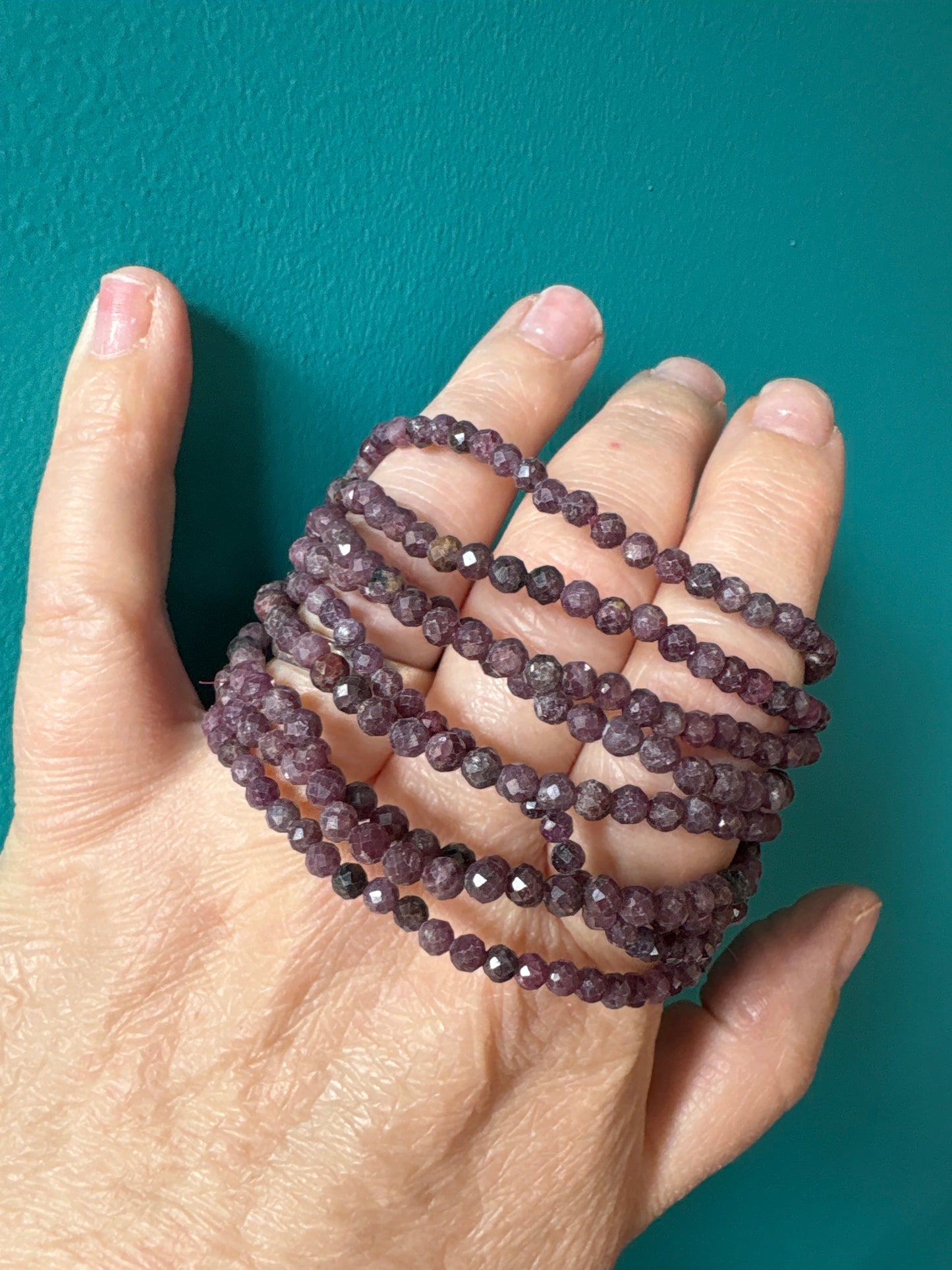 Close-up of hand holding multiple strands of faceted 4mm deep crimson ruby bracelets against teal background