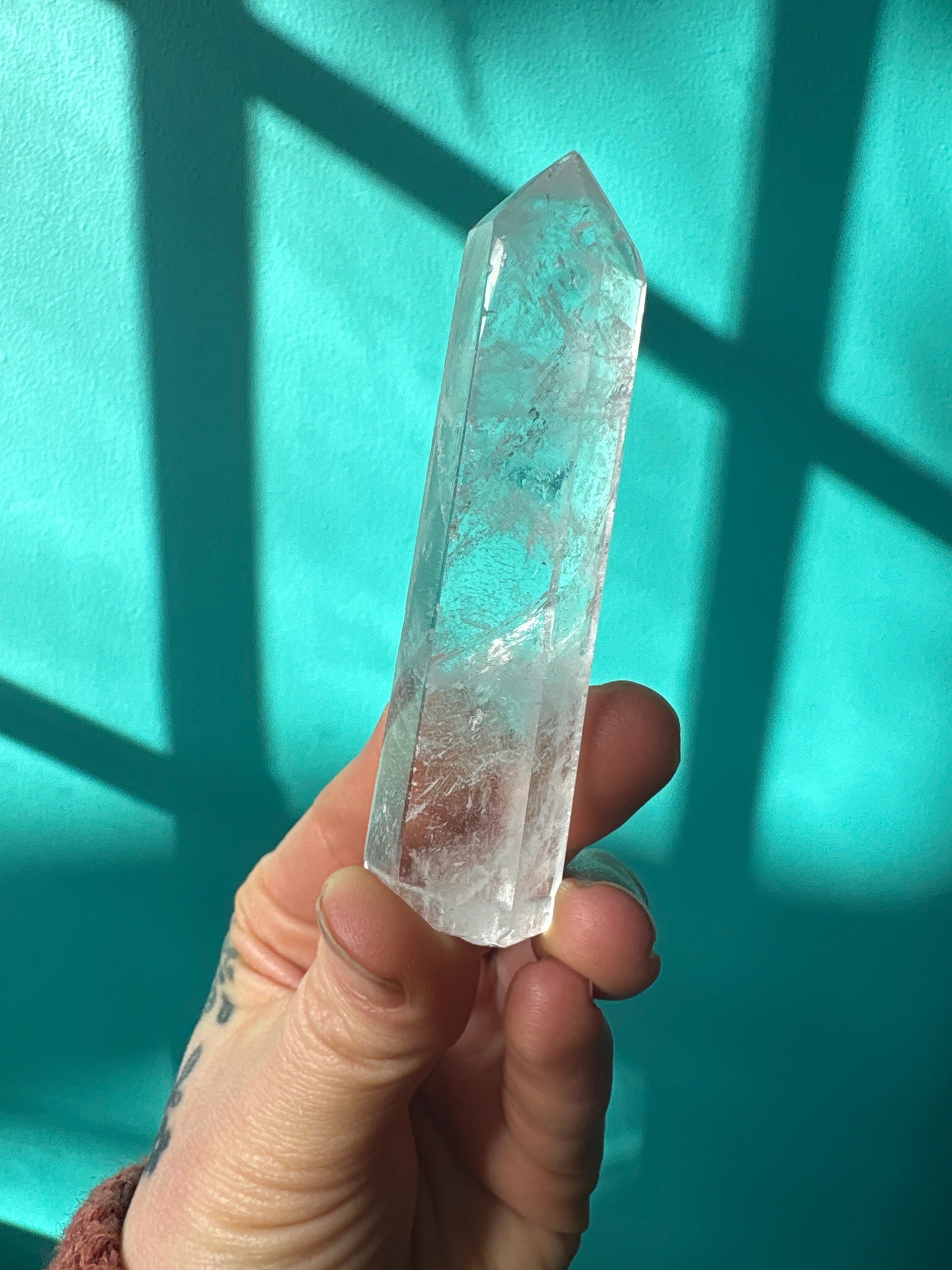 Hand holding a 3-4 inch clear quartz tower crystal against a turquoise background