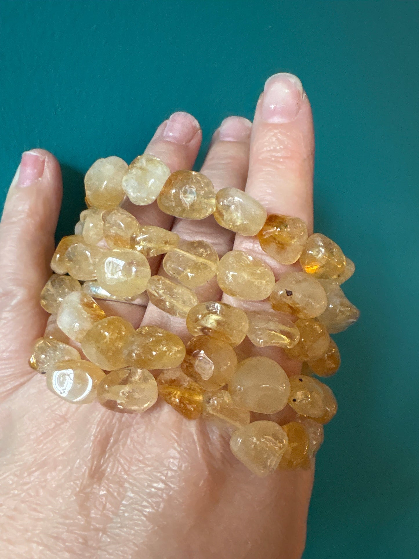 Hand holding multiple strands of tumbled Citrine gemstone bracelets 8-10mm beads against teal background