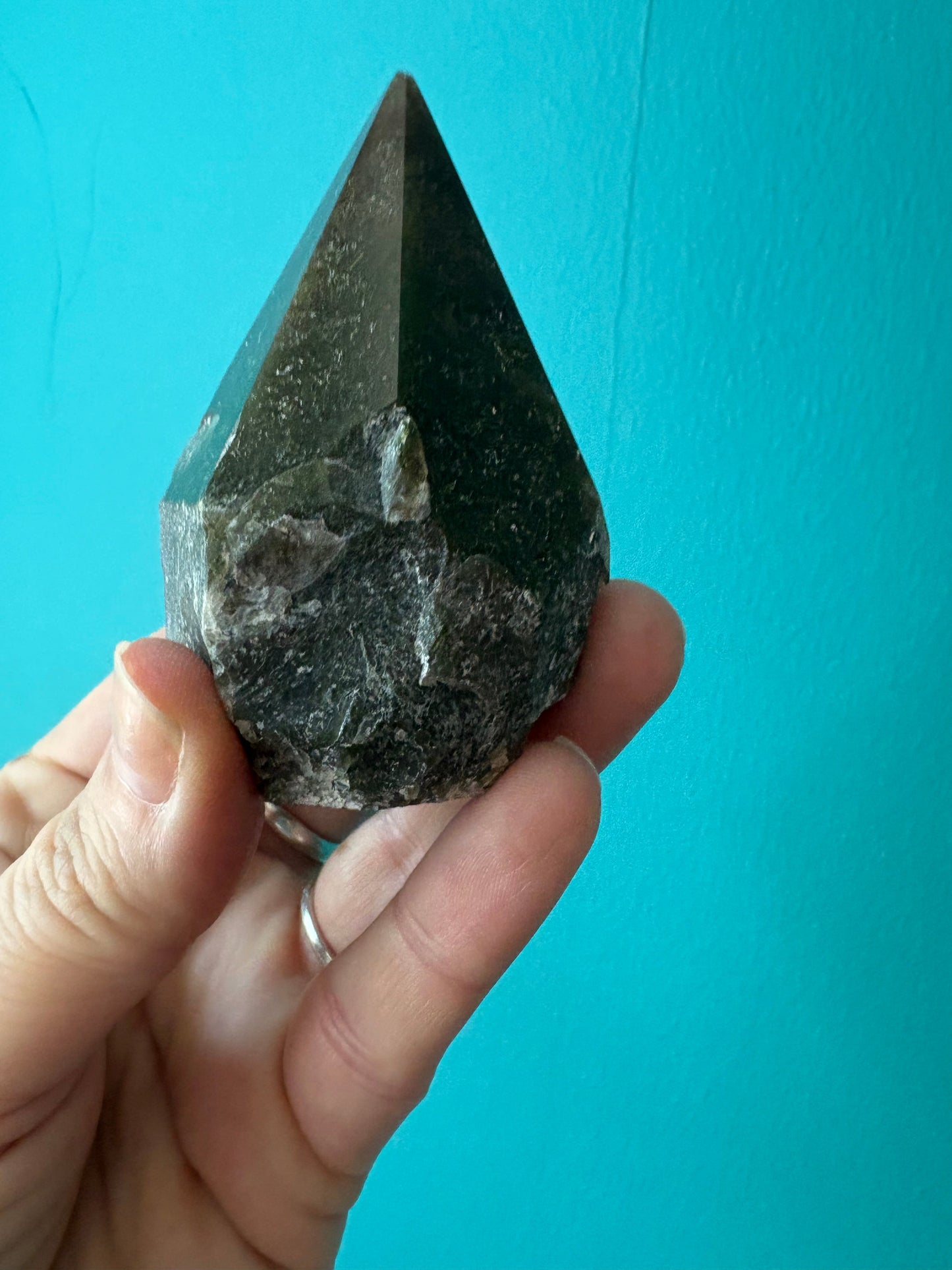 Hand holding a deep green Moss Agate point crystal with moss-like inclusions against a turquoise background