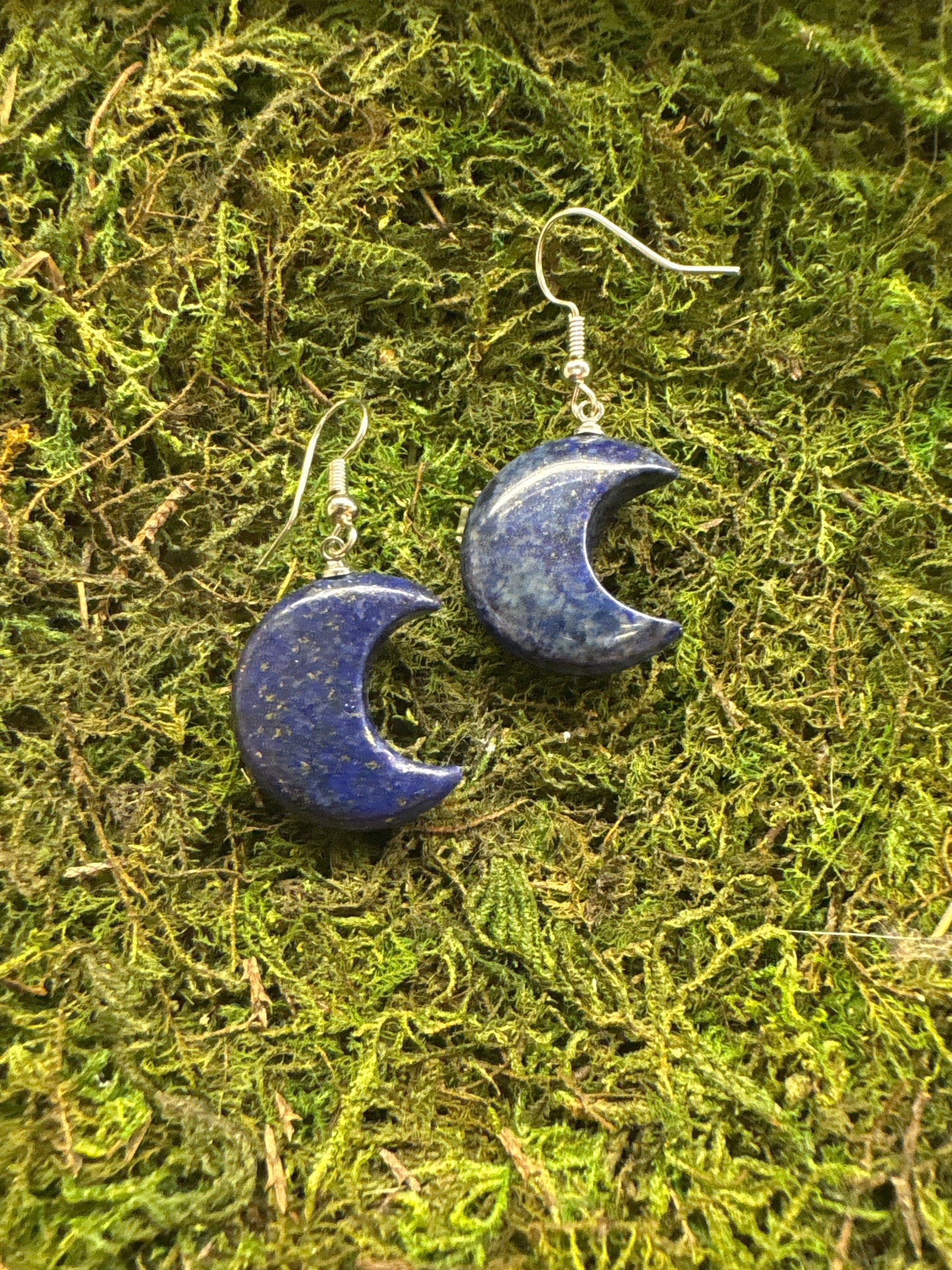Lapis Lazuli moon-shaped earrings with deep blue hues and silver hooks on green moss background