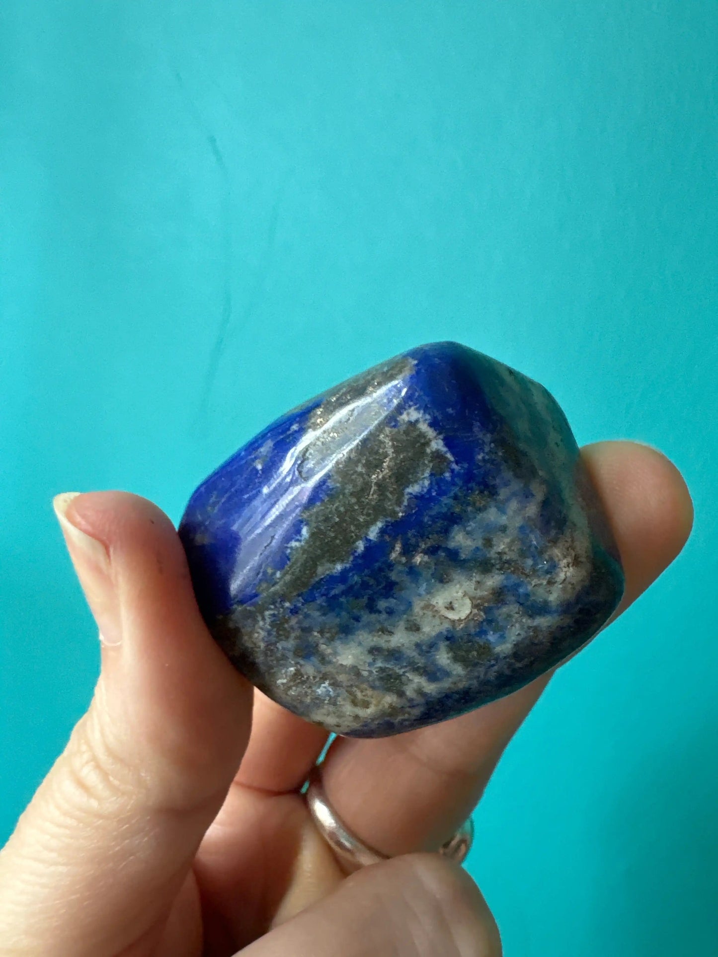 hand holding a polished tumble lapis lazuli stone with blue and gray tones against a turquoise background