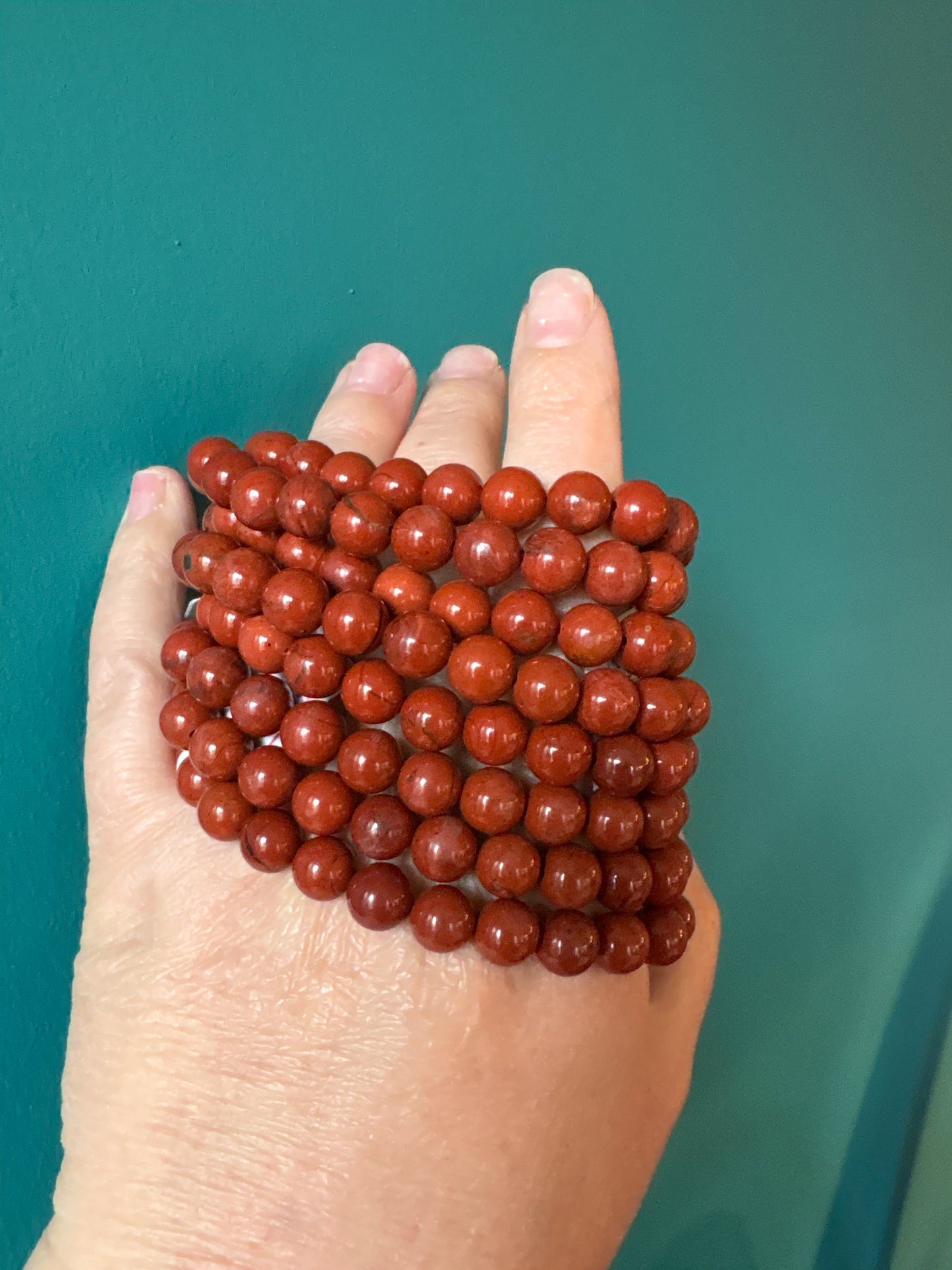 Close-up of hand holding multiple strands of 8mm red jasper bead bracelets against a teal background