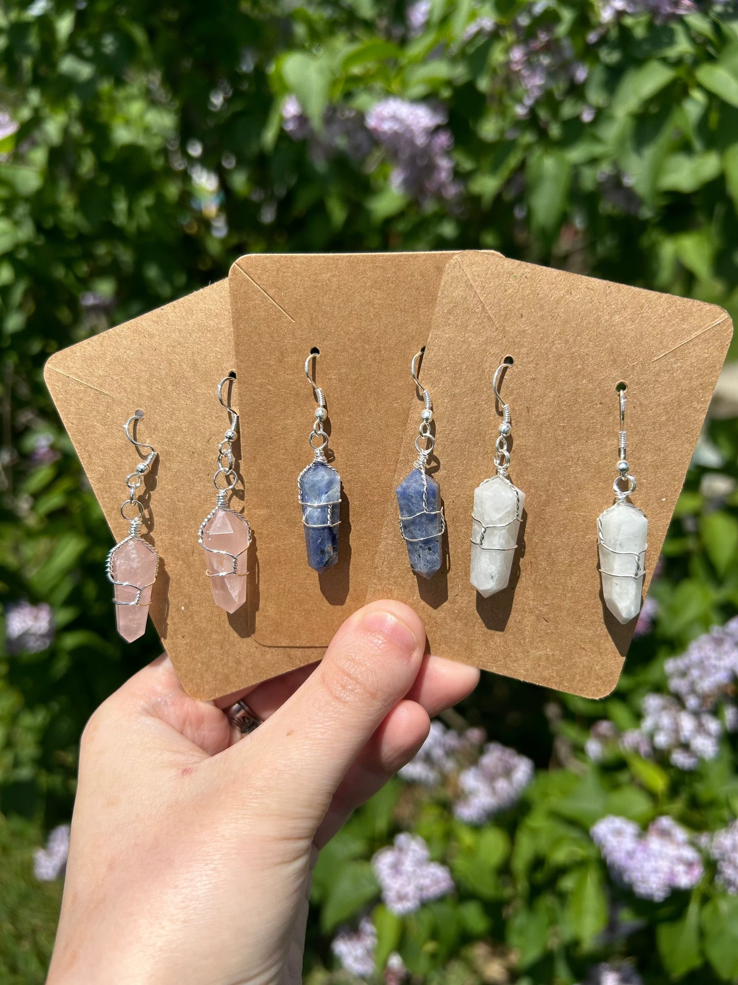 Handcrafted wire wrap crystal earrings in pink, blue, and white displayed on brown cards outdoors with greenery background
