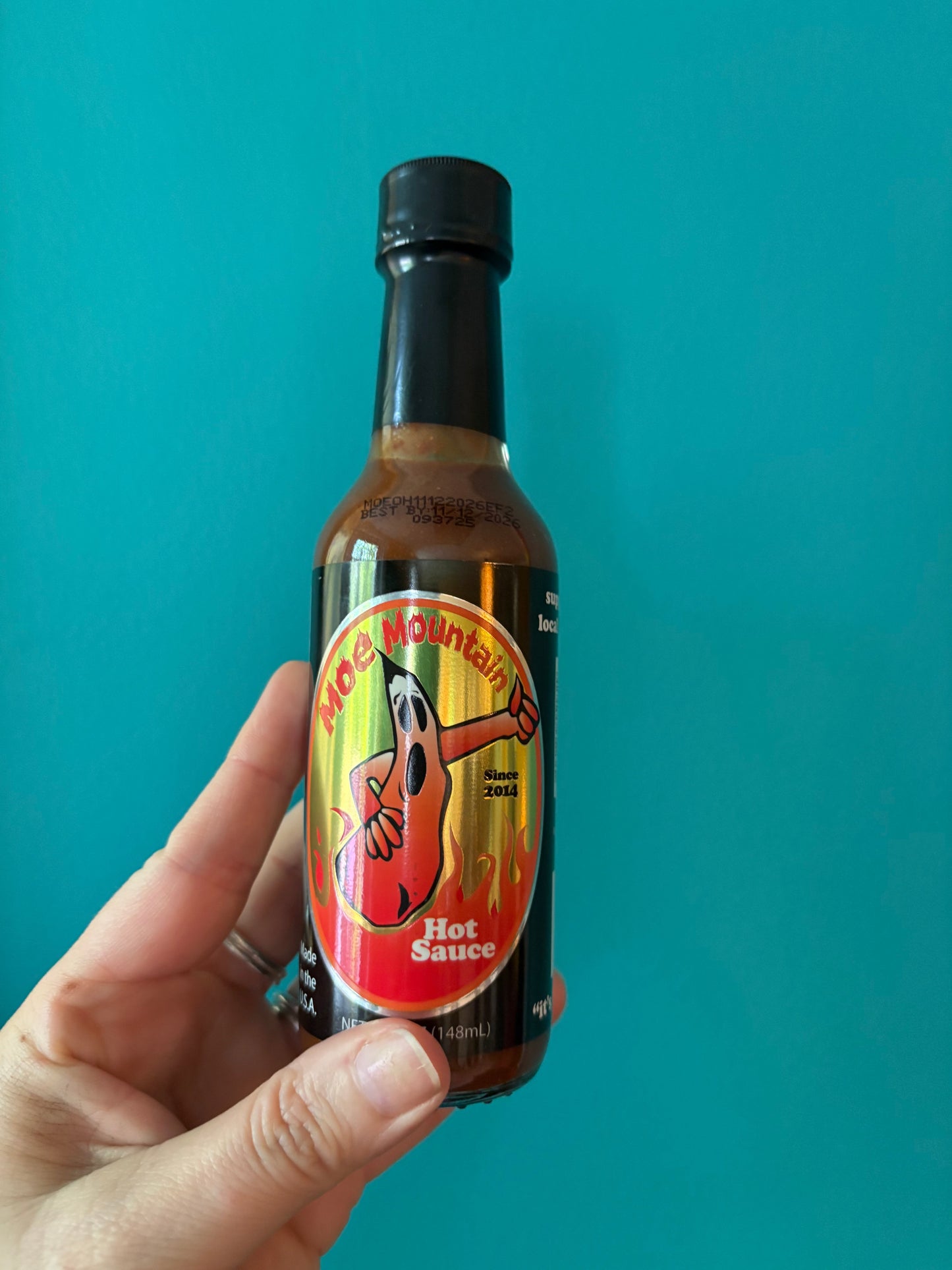 Bottle of Moe Mountain Hot Sauce with spicy pepper label held against blue background