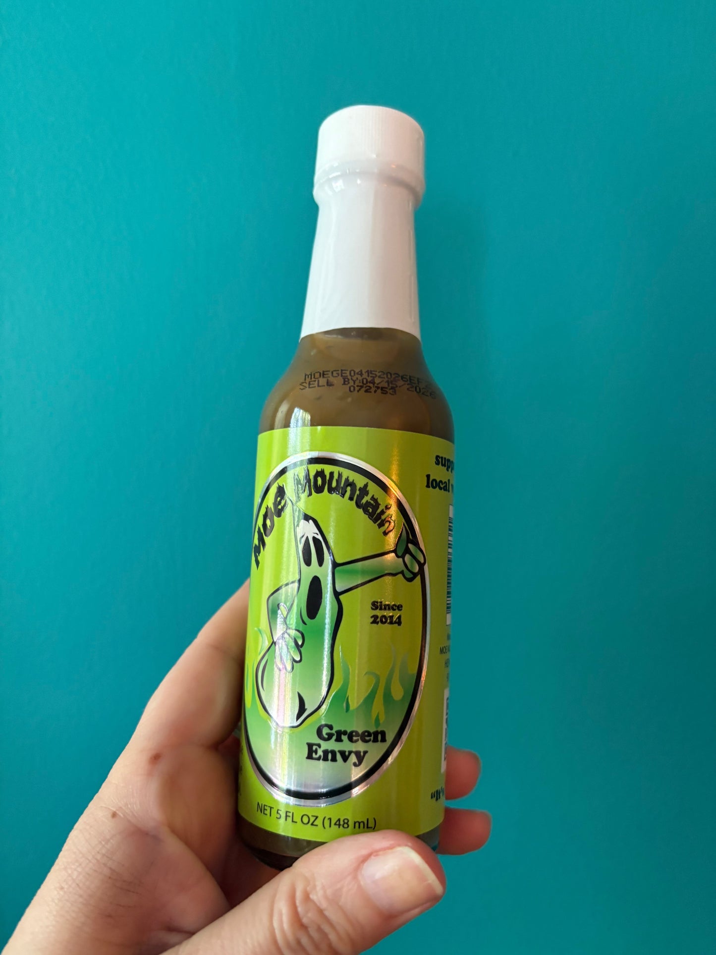 Hand holding Moe Mountain Green Envy hot sauce bottle with green label against blue background
