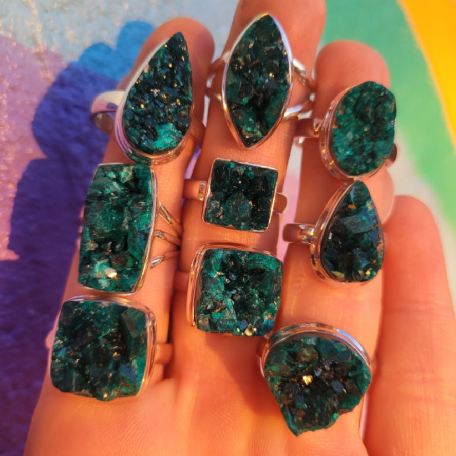 Hand displaying multiple Dioptase Druzy rings with vibrant green-blue crystalline stones in various shapes under sunlight