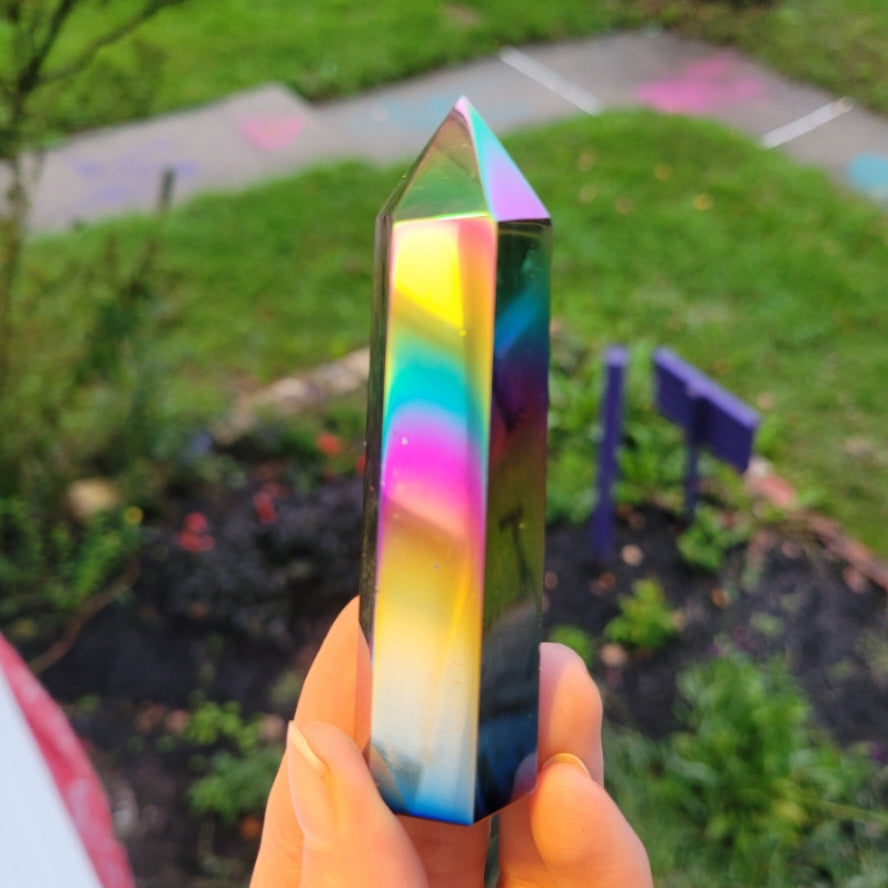 Hand holding a colorful Obsidian Angel Aura Tower crystal outdoors with blurred greenery background