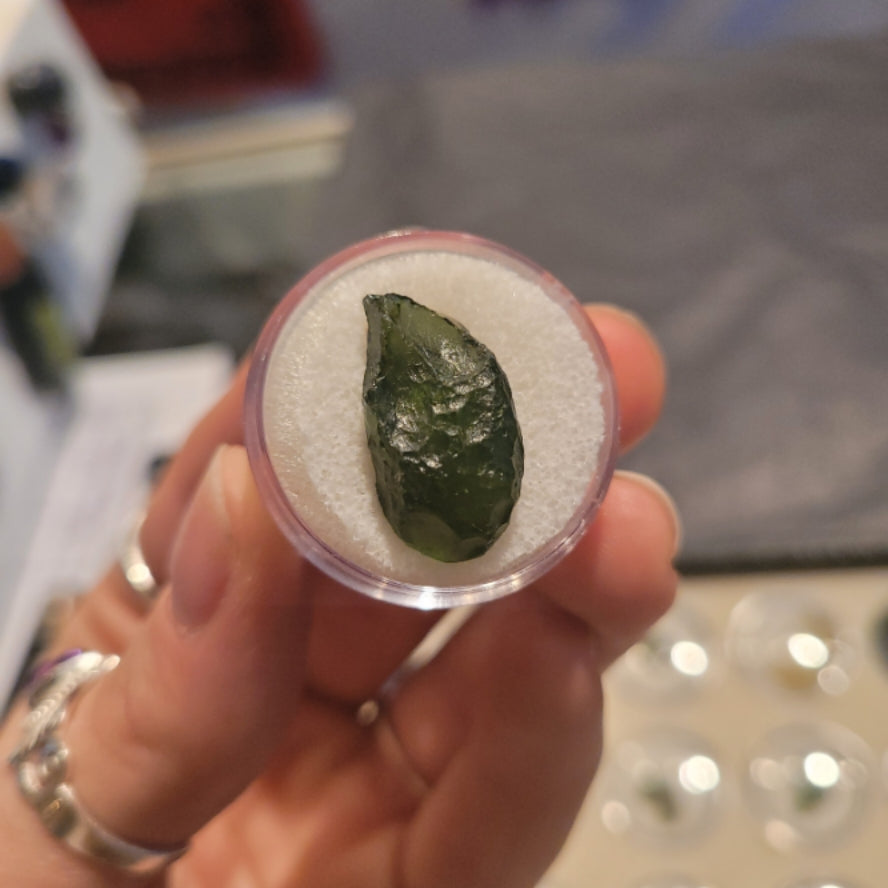 Close-up of a green moldavite tektite from the Czech Republic held in a small clear container with a white cushion inside.