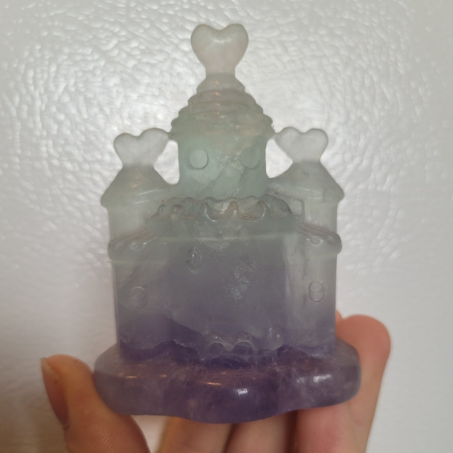 Hand holding a purple and green Castle Fluorite crystal carved in the shape of a fluorite house with heart-shaped turrets.