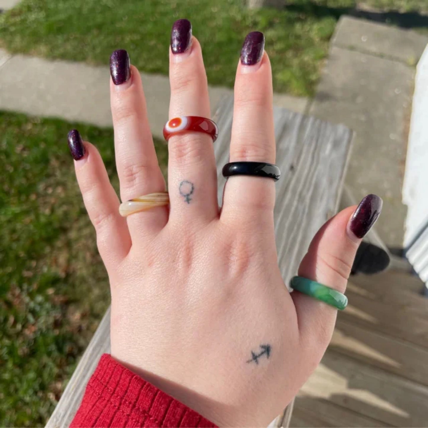 Hand wearing mixed agate and jasper rings in varying colors outdoors with purple nail polish and small tattoos on fingers