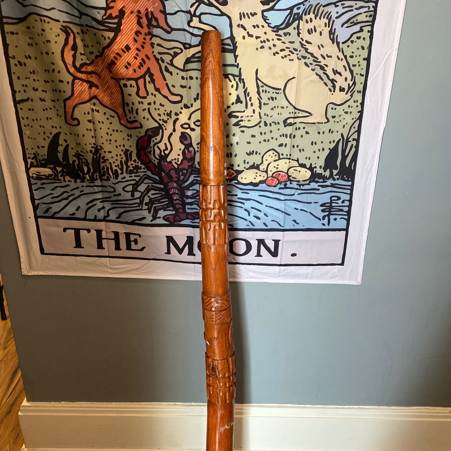 Wooden didgeridoo standing upright with carved details against a wall with decorative tapestry background
