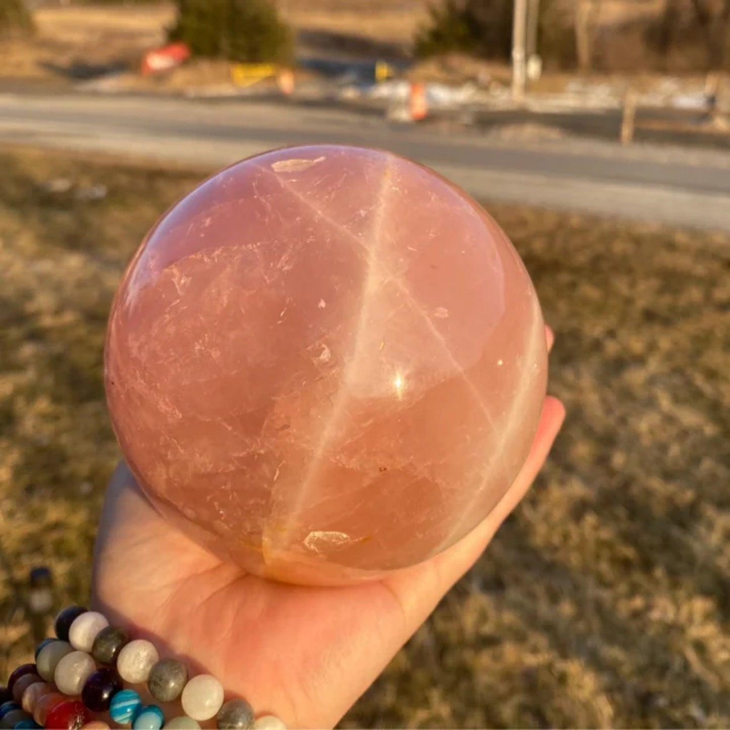 Hand holding a polished natural rose quartz sphere reflecting sunlight outdoors with a blurred grassy background