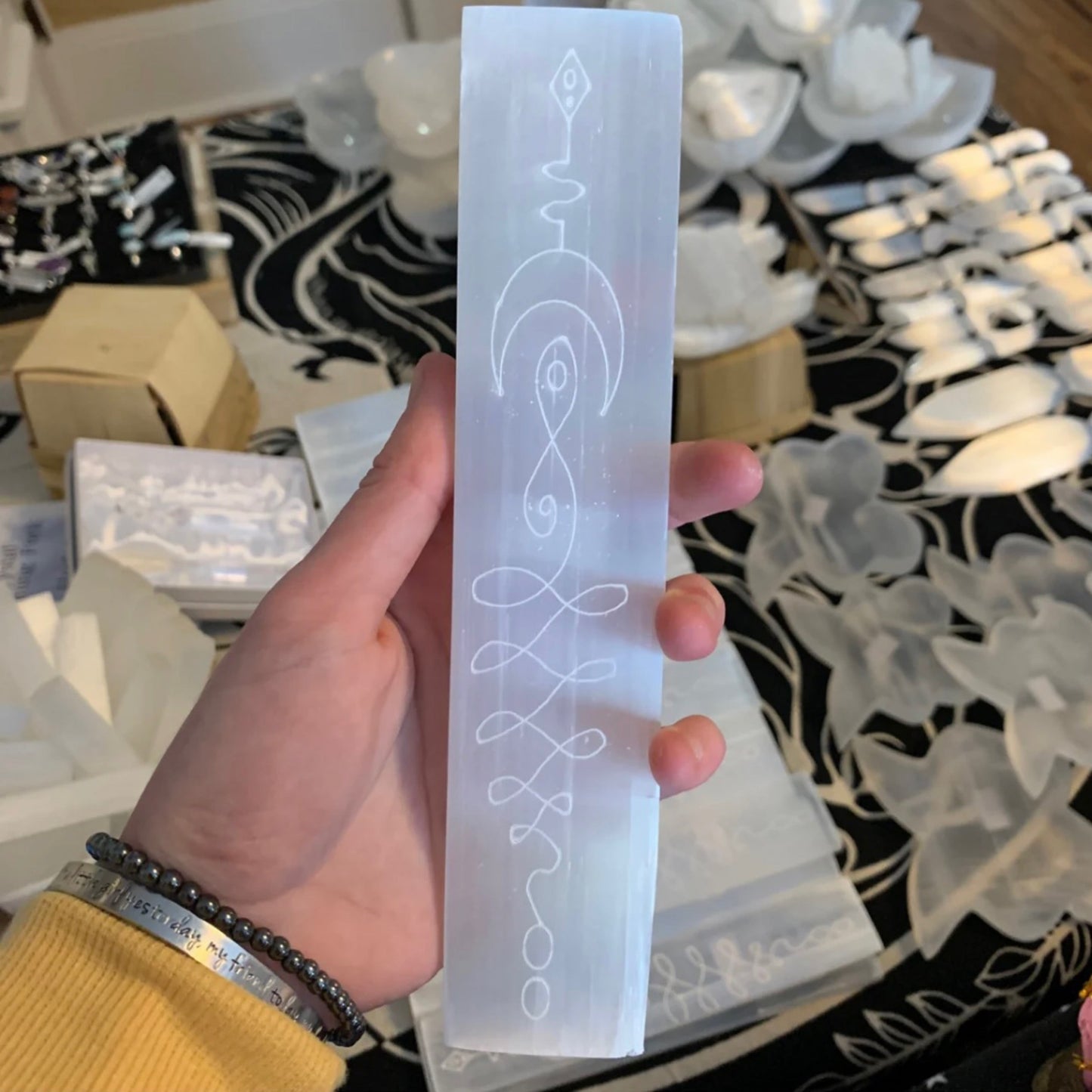 Hand holding a rectangular selenite moon plate with engraved mystical symbols, surrounded by various crystal decor pieces in the background