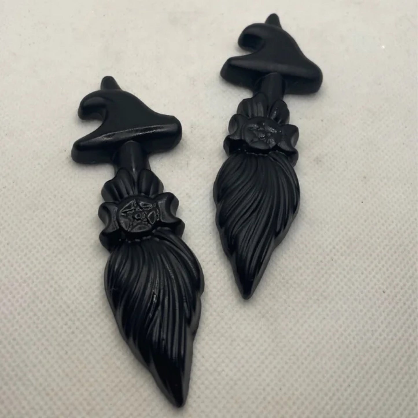 Two black obsidian broom-shaped figurines with witch hats and floral details on a white background
