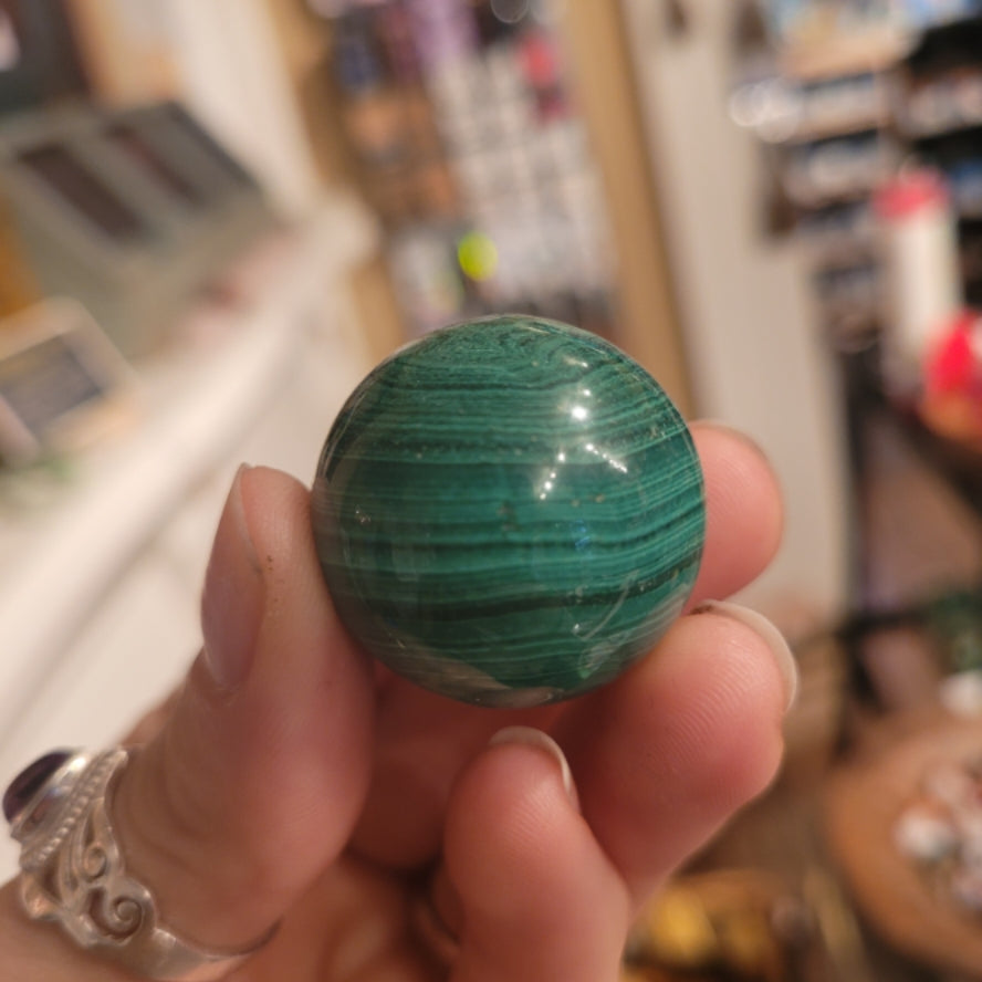 Hand holding a medium malachite sphere with green banded patterns in an indoor setting