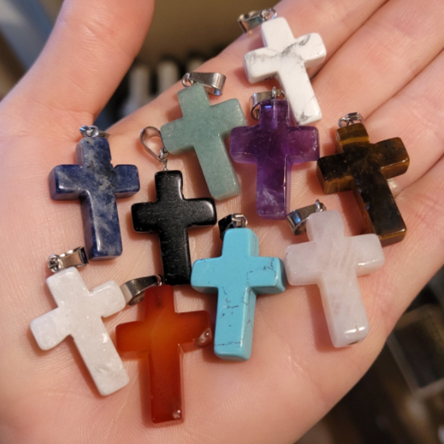 Hand holding assorted colorful crystal cross pendants including carnelian, amethyst, turquoise, and marble variations
