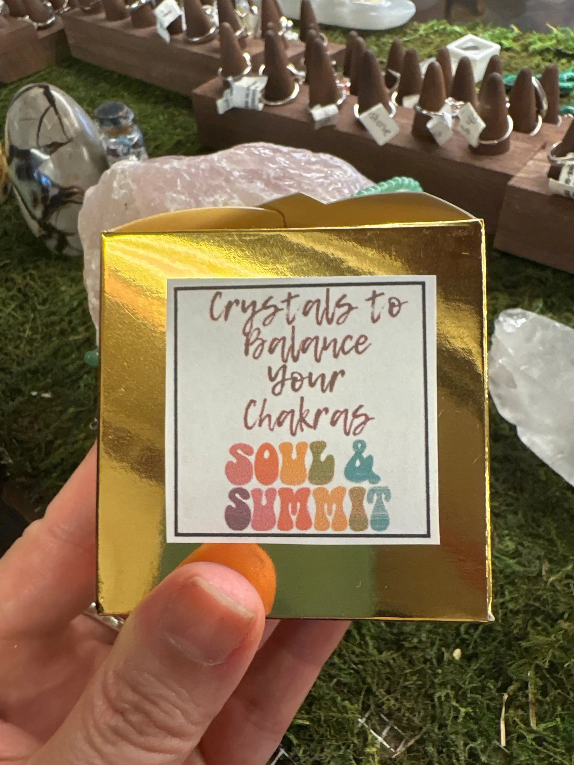 Golden box labeled "Crystals to Balance Your Chakras Soul & Summit" held in hand outdoors