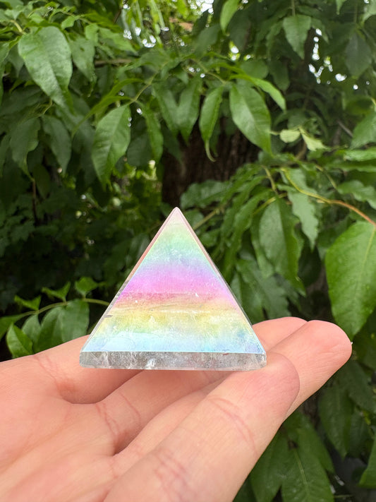 Hand holding a shimmering Angel Aura Quartz Pyramid with iridescent rainbow colors against green foliage background