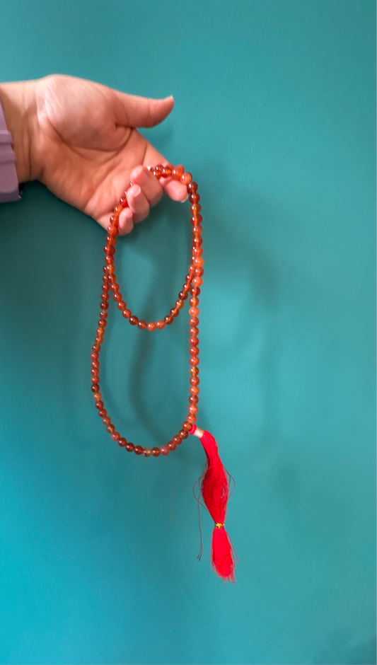 Hand holding a Carnelian Bead Mala with 108 orange-red beads and a red tassel against a turquoise background