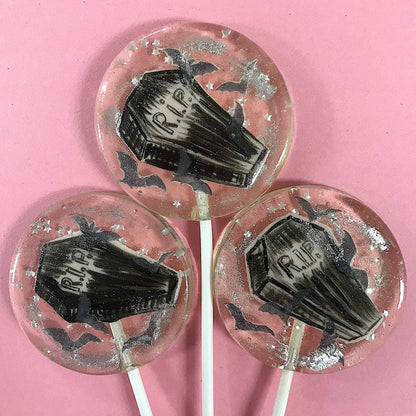 Round lollipops with black fondant coffin designs, silver glitter stars, and wafer paper bats on a pink background Peace Coffin Lollipops