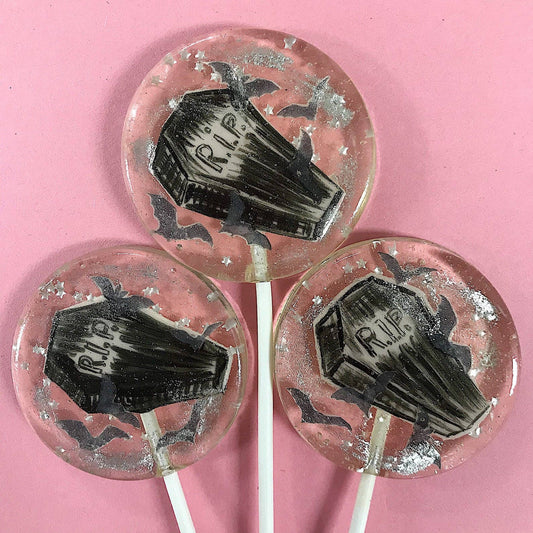 Round lollipops with black fondant coffin designs, silver glitter stars, and wafer paper bats on a pink background Peace Coffin Lollipops