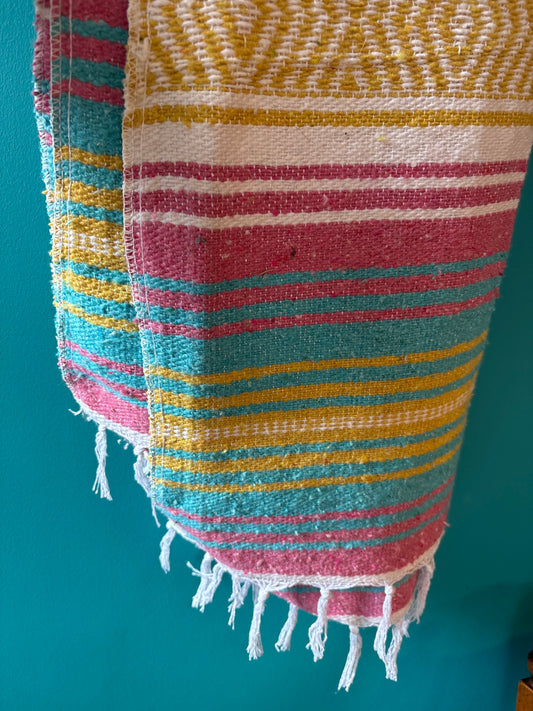 Handwoven vibrant Mexican blanket with colorful stripes and fringed edges hanging against a teal wall