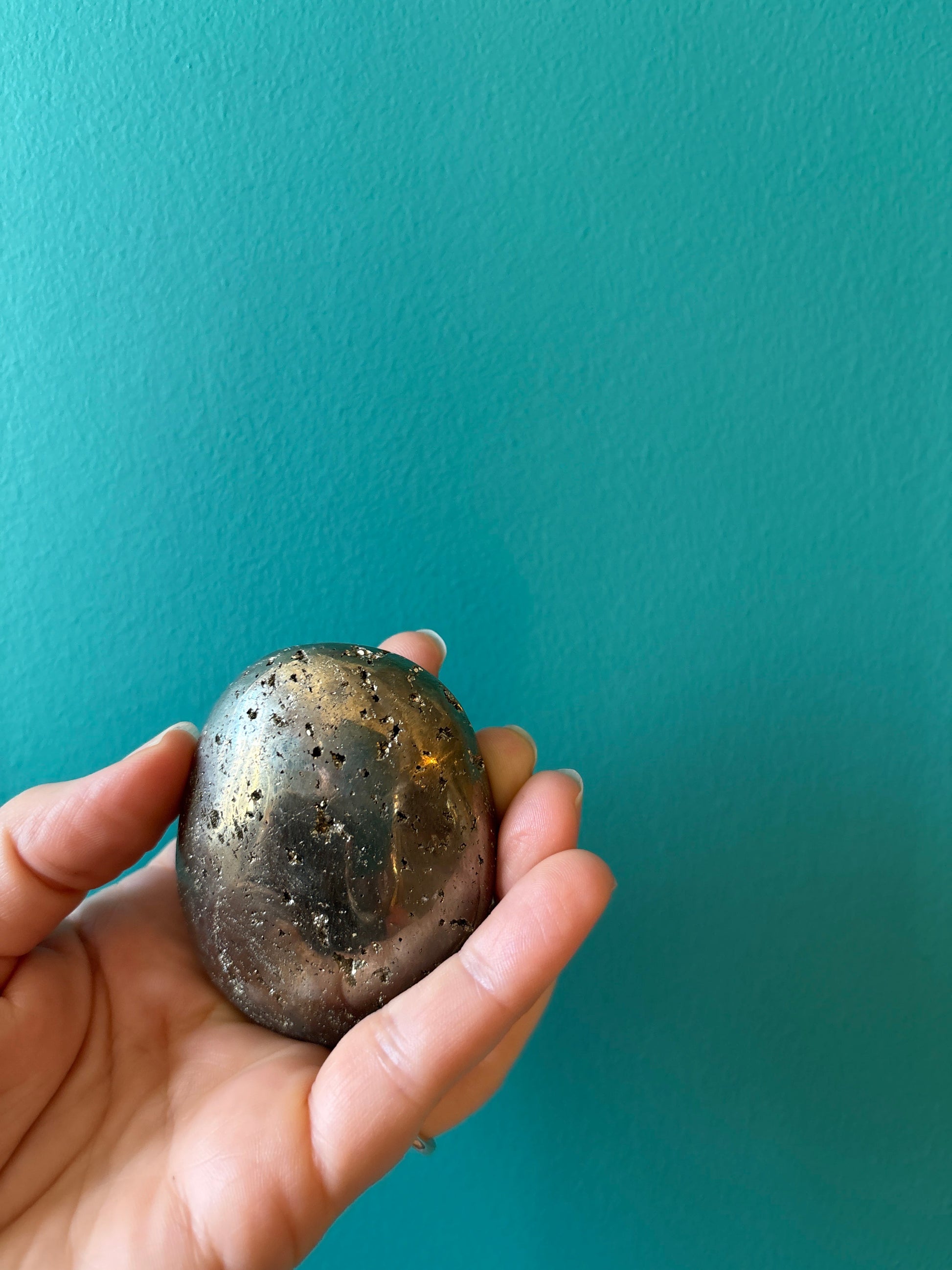 Hand holding a shiny pyrite palm stone against a teal background, symbolizing prosperity and abundance.