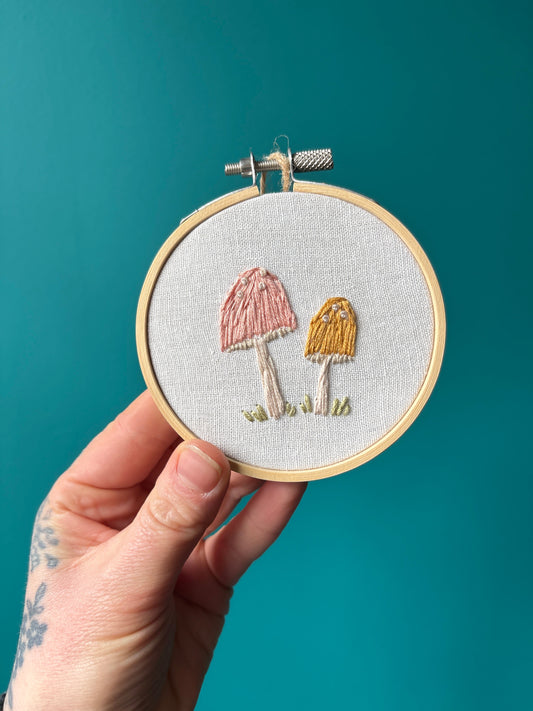 Handmade embroidery of two mushrooms in a wooden hoop on white fabric against teal background