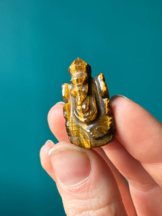 Handcrafted crystal mini Ganesh statue carved from tiger's eye held between fingers against a teal background