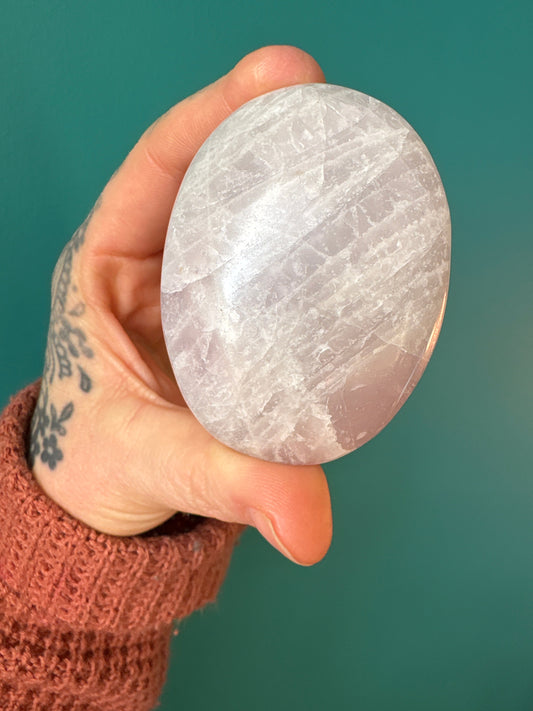 Hand holding a polished Kunzite palm stone against a teal background