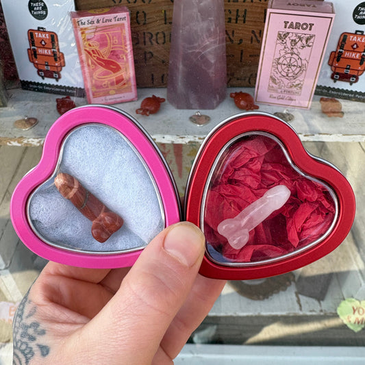 Two crystal weenie figurines in separate metal heart boxes, one pink and one red, held in a hand with tarot cards in the background