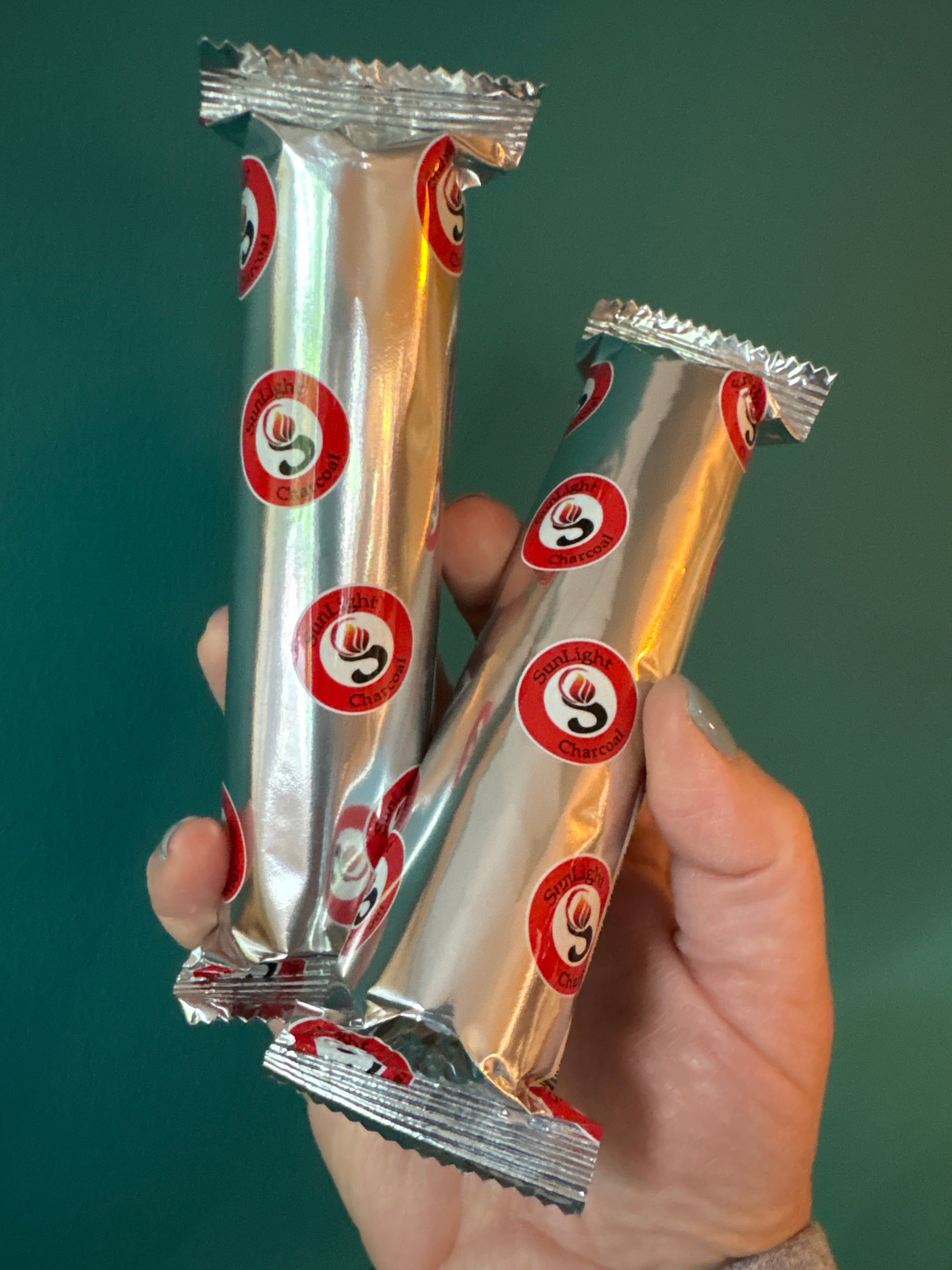 Hand holding two silver foil packs of charcoal tablets with red and white circular logos on a green background