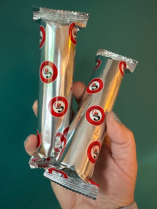 Hand holding two silver foil packs of charcoal tablets with red and white circular logos on a green background